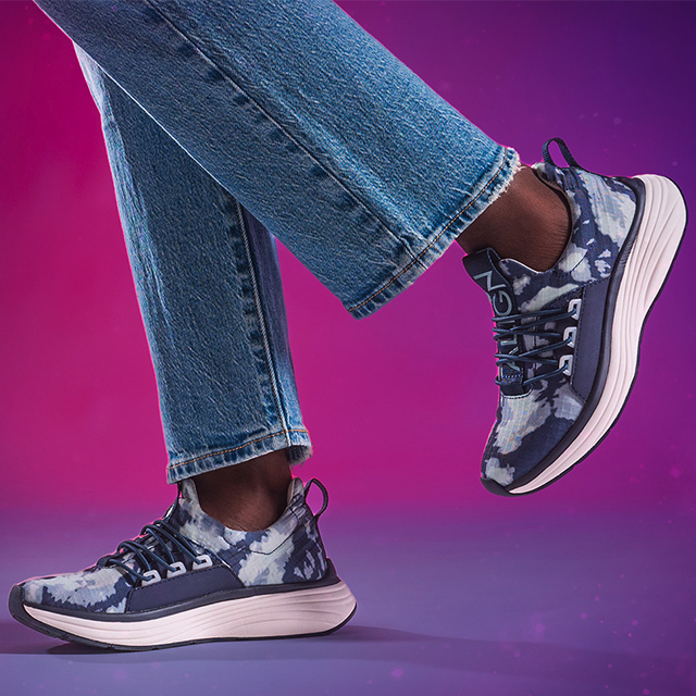Women’s Theora Sneakers in Blue Tie Dye on foot on a pink and purple background with the back foot in the air